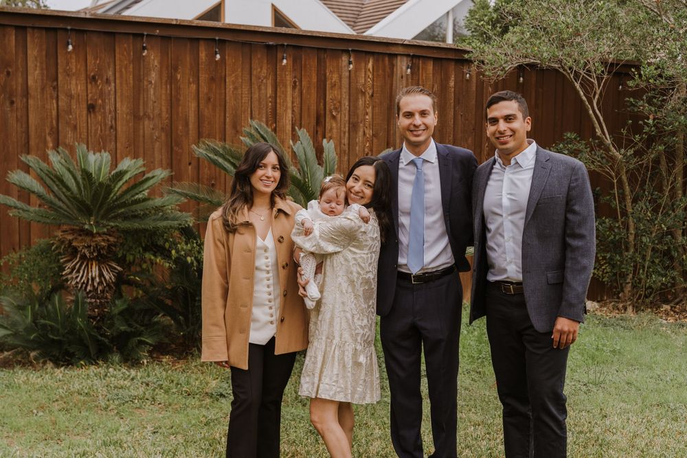 Small group poses together with infant in formal attire against wooden fence and landscaping.