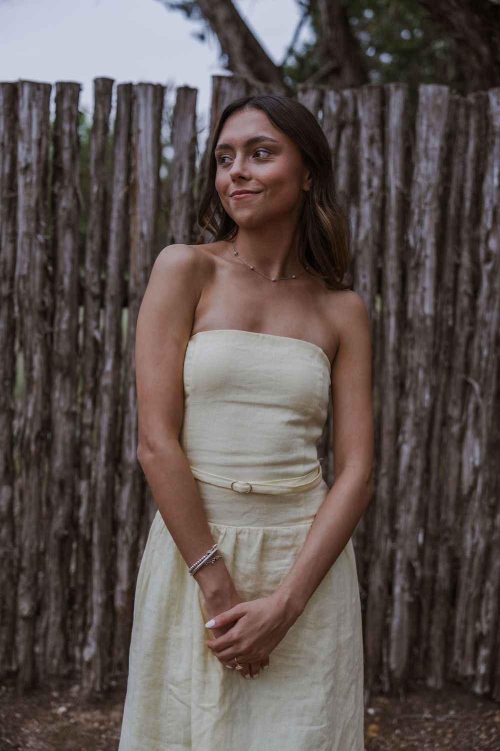 A woman in a white strapless dress poses against a rustic wooden fence backdrop.