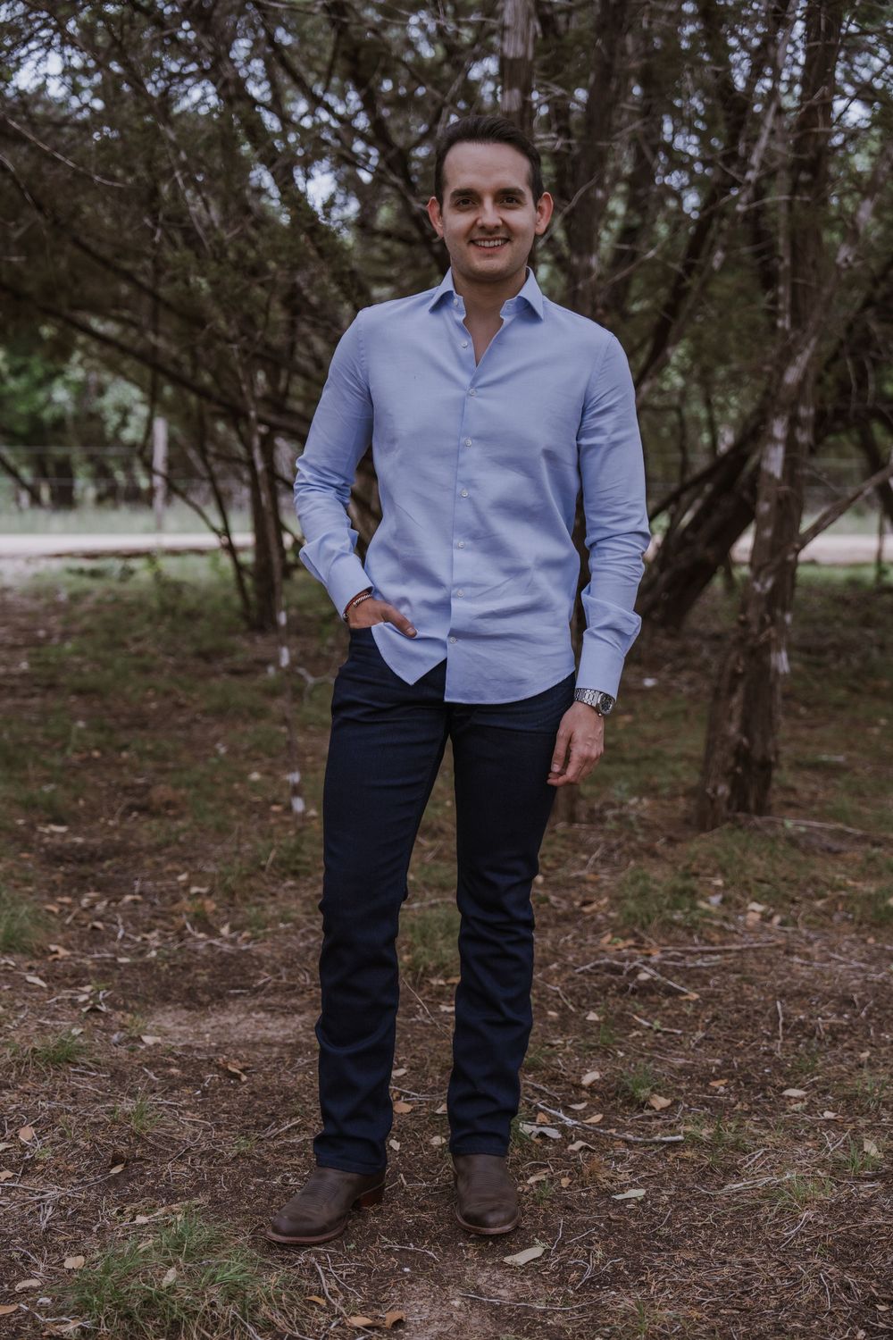 A person in a light blue button-down shirt stands among trees in a wooded setting.