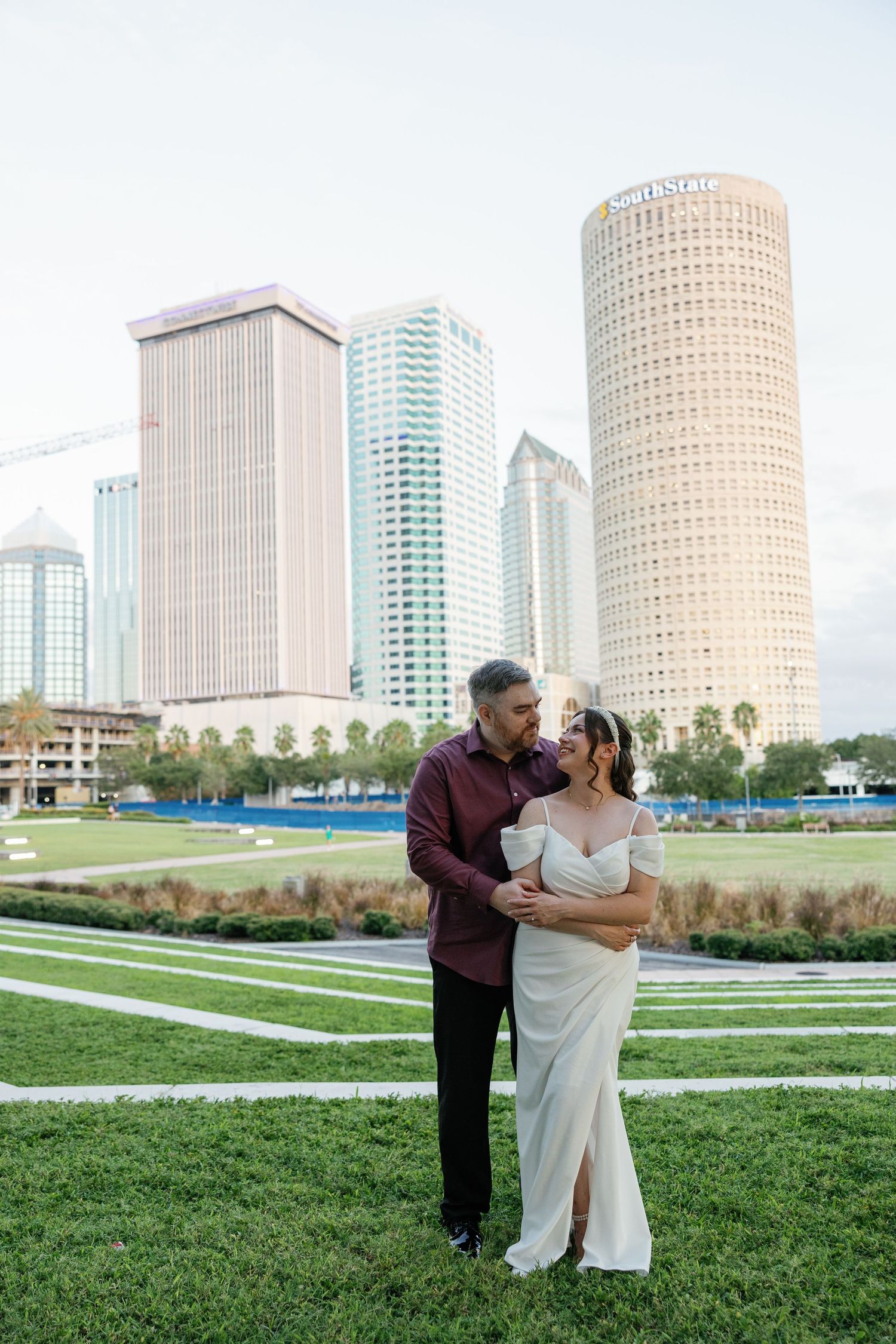 A couple in wedding attire playfully poses on an urban street with modern buildings in the background.