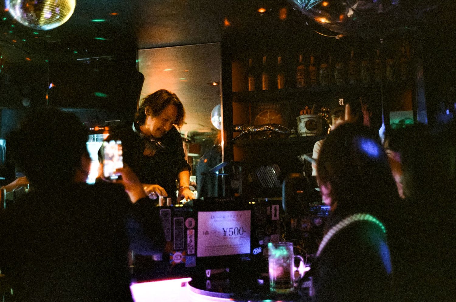 A DJ performs at a dimly lit nightclub with disco balls overhead and colorful lights in the background.
