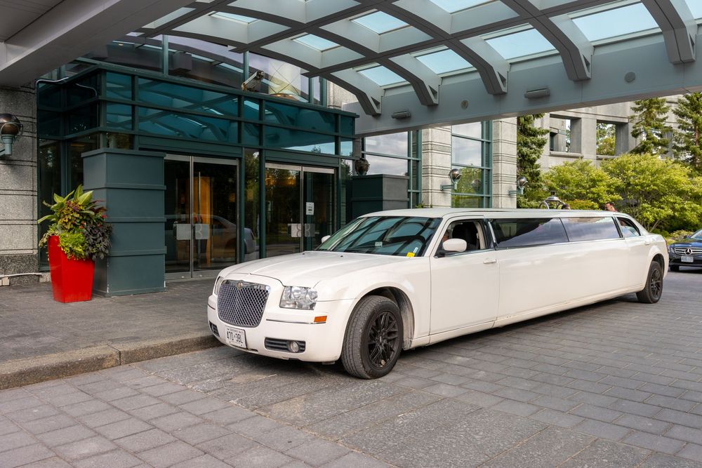White Chrysler 300 stretch limousine parked at entrance of modern building with glass canopy and red planter.
