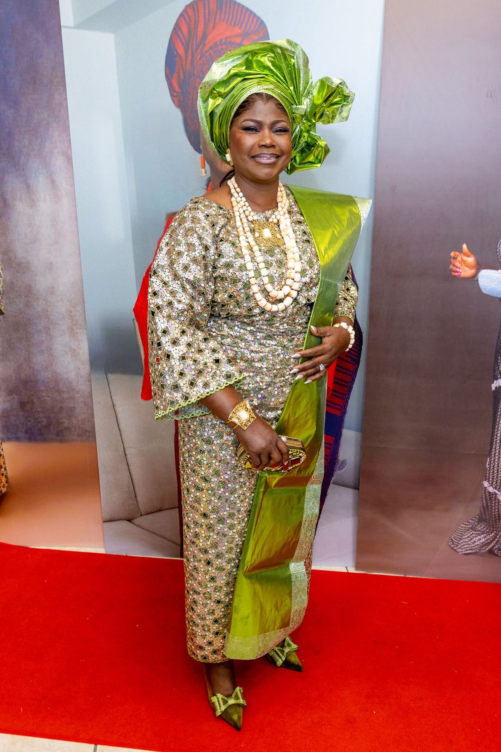 Traditional Nigerian attire featuring a patterned dress, green headwrap, and pearl necklaces on a red carpet.