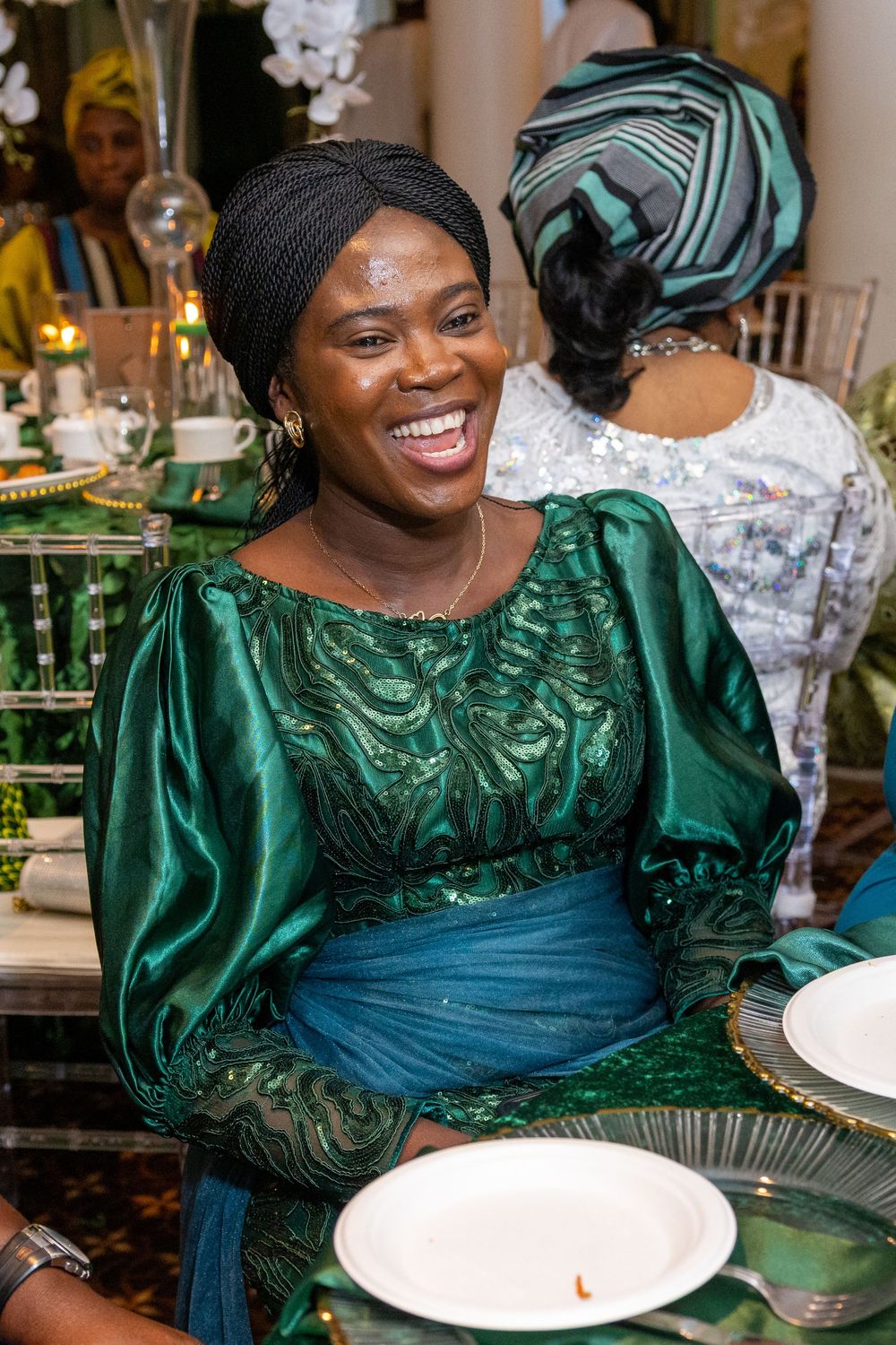 A person in an emerald green traditional dress with ornate details sits at a formal dining table smiling.