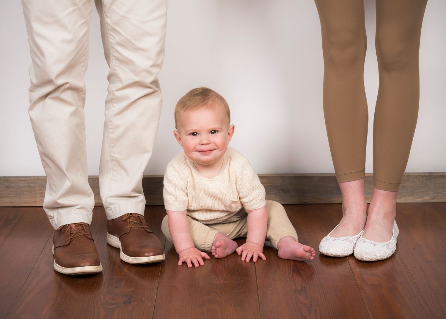 A series of feet and legs standing on hardwood flooring with a small child crawling between them.