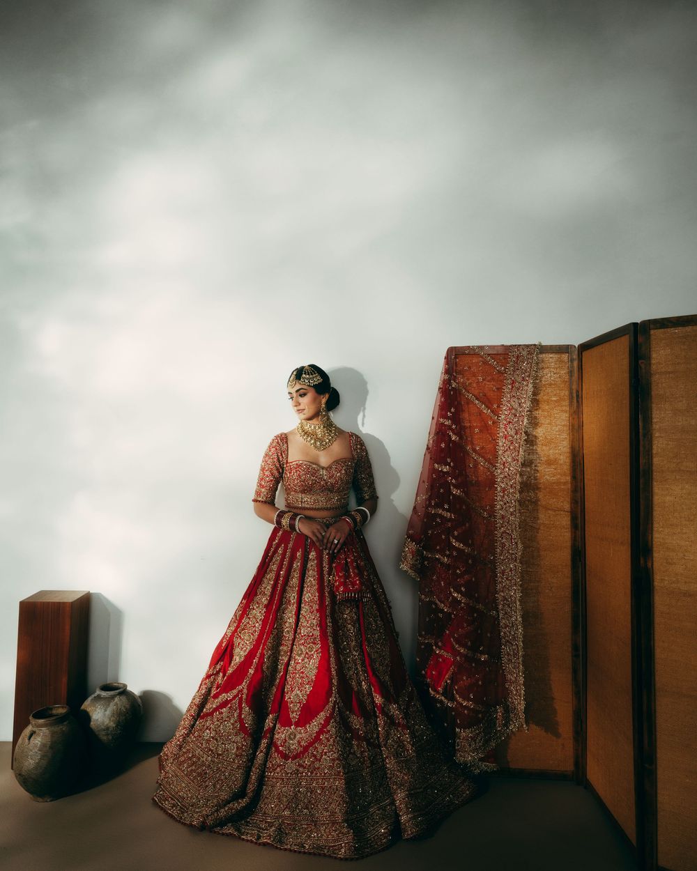 A traditional red and gold Indian bridal lehenga dress displayed against a dark moody backdrop with dramatic lighting.