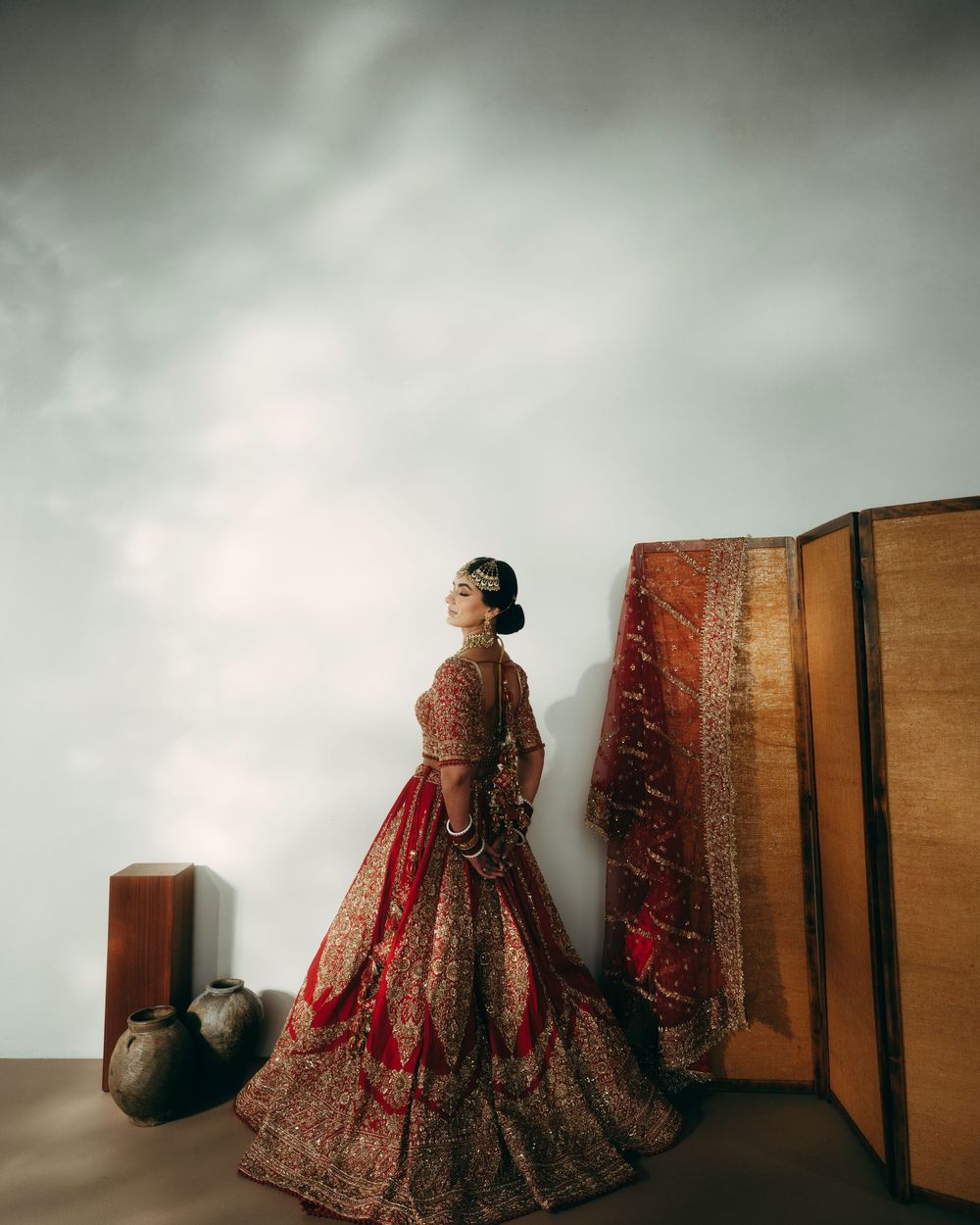 A figure in a traditional red and gold embroidered wedding lehenga sits elegantly against a wooden backdrop.