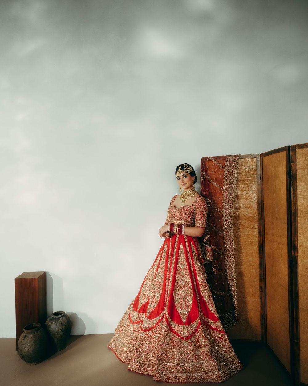 Elegant red and gold bridal lehenga showcased in artistic poses against minimalist backdrop with wooden panels.