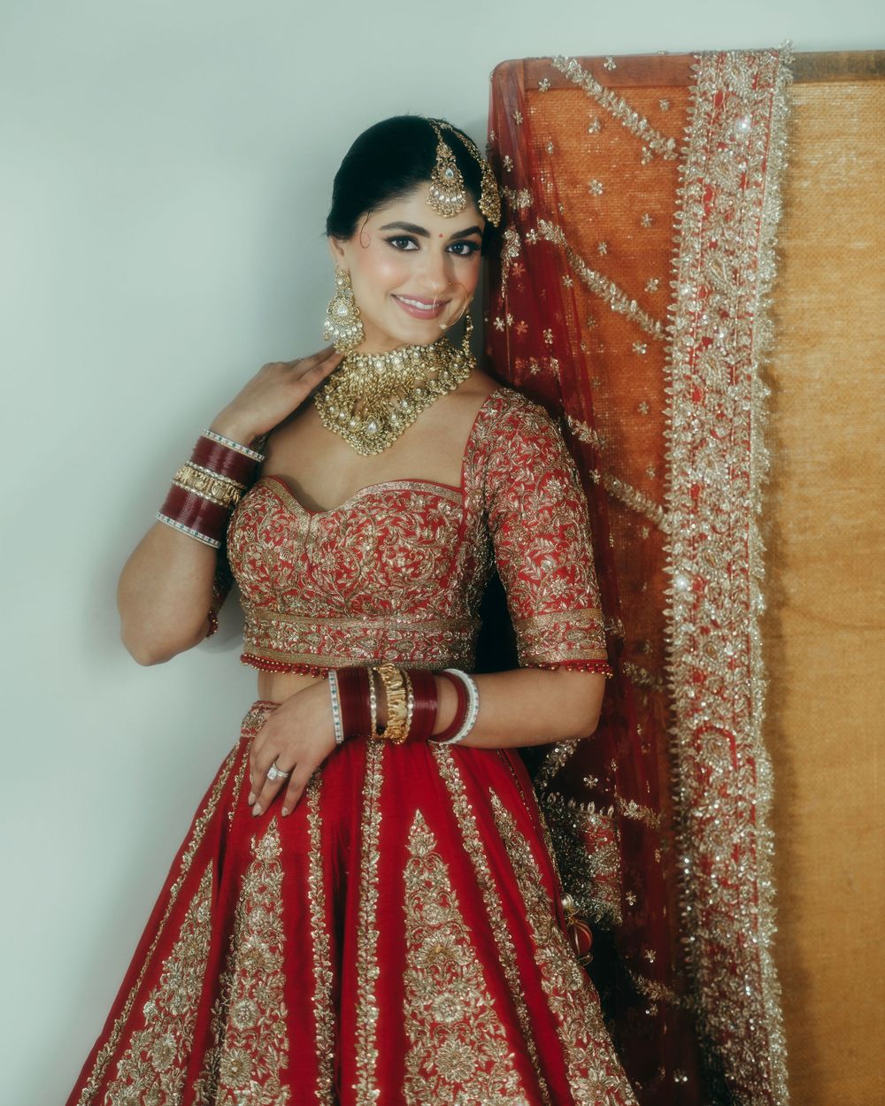A person wearing a traditional red and gold embroidered Indian bridal lehenga poses elegantly against a light background.