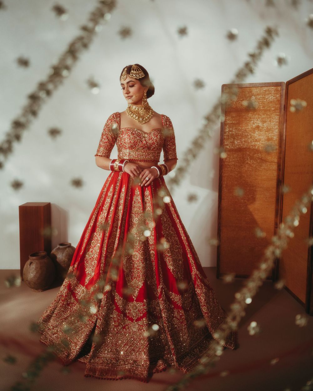 Traditional red and gold embroidered bridal lehenga photographed in artistic sequence against white wall with bokeh lights.