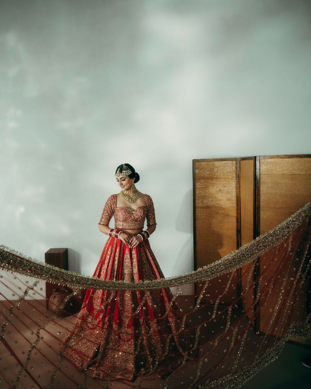 An elegant bridal pose showcasing a flowing red and gold lehenga with intricate embroidery against wooden panels.