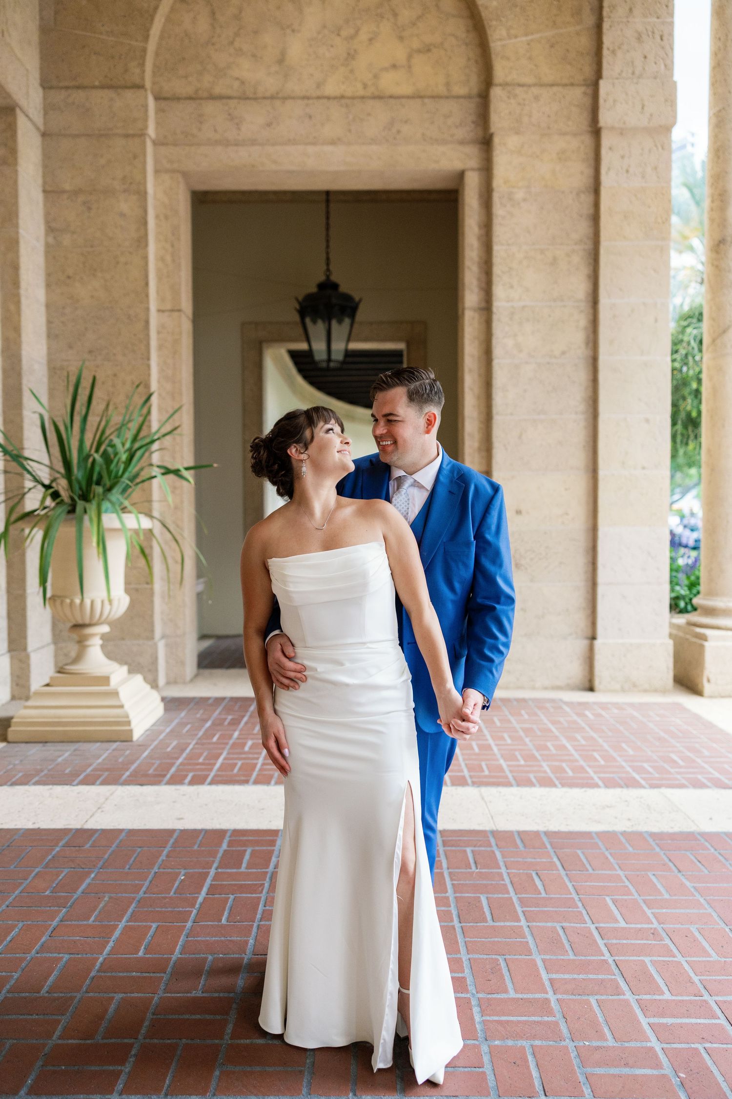 Romantic couple sits on a stone wall overlooking a waterfront path lined with palm trees and oak branches.