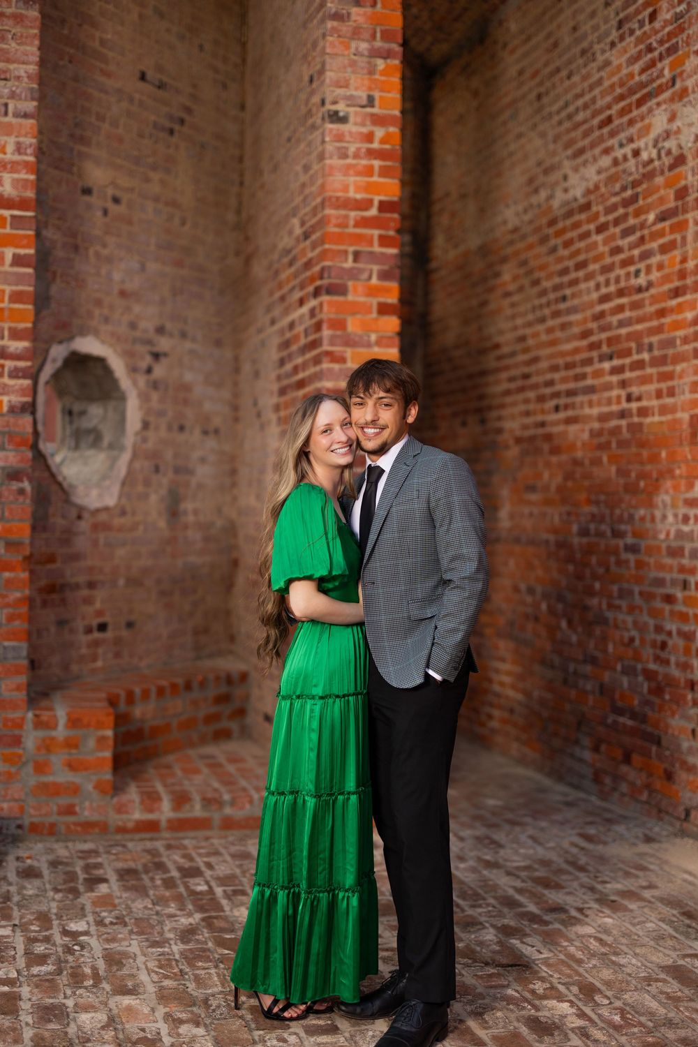 Couple poses in romantic engagement photo against rustic brick wall interior.