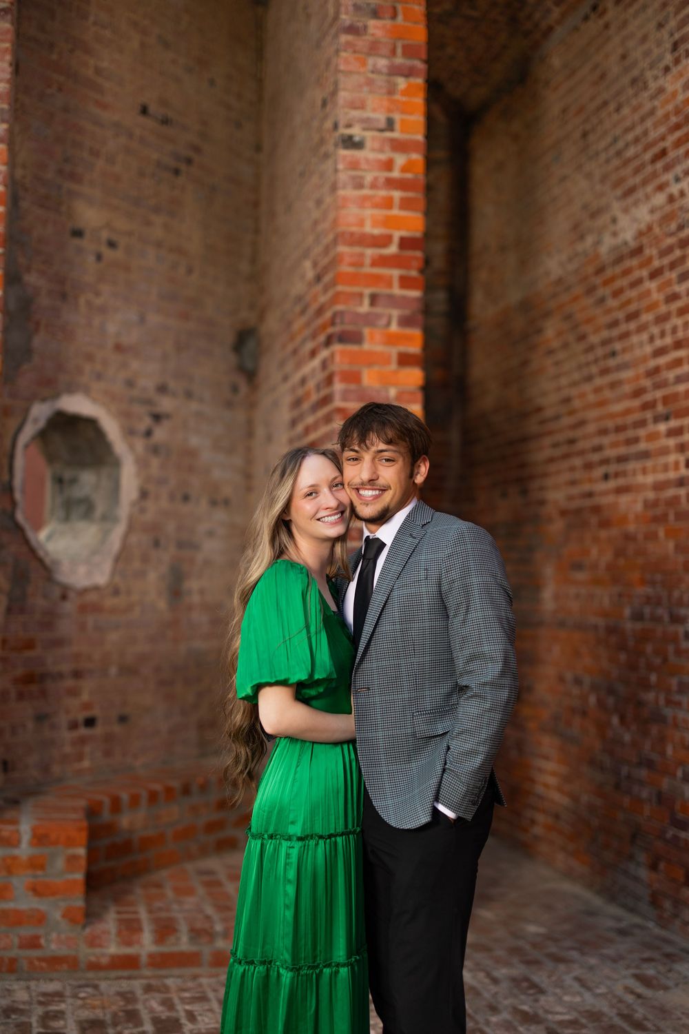 Couple in formal wear embracing against rustic brick wall background.