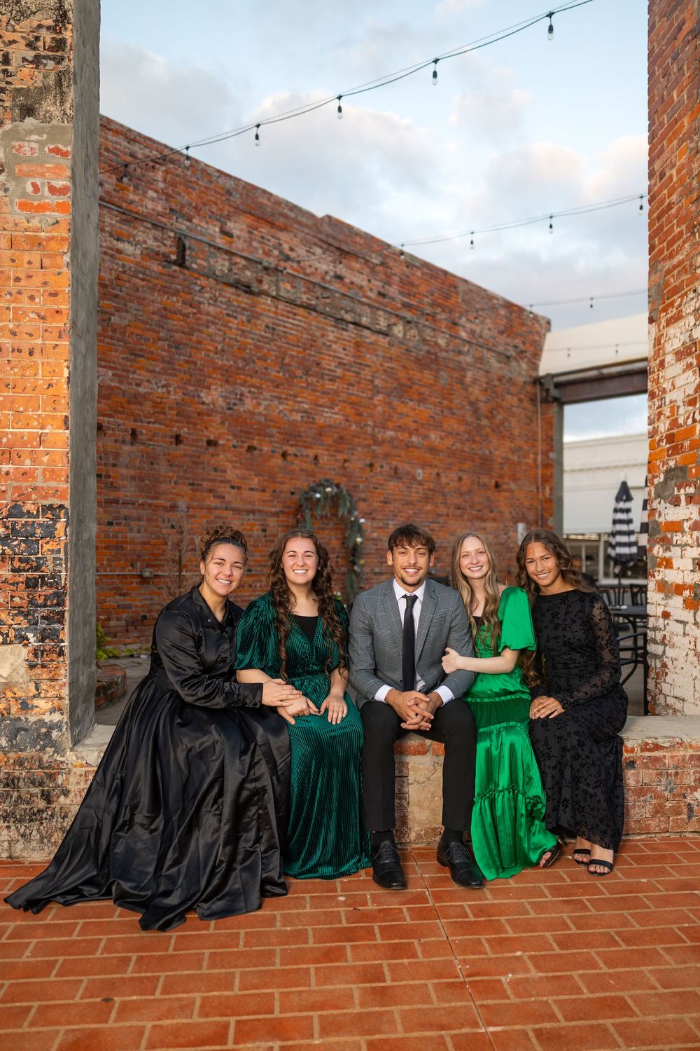 Group photo with five people in formal attire against brick wall with string lights.