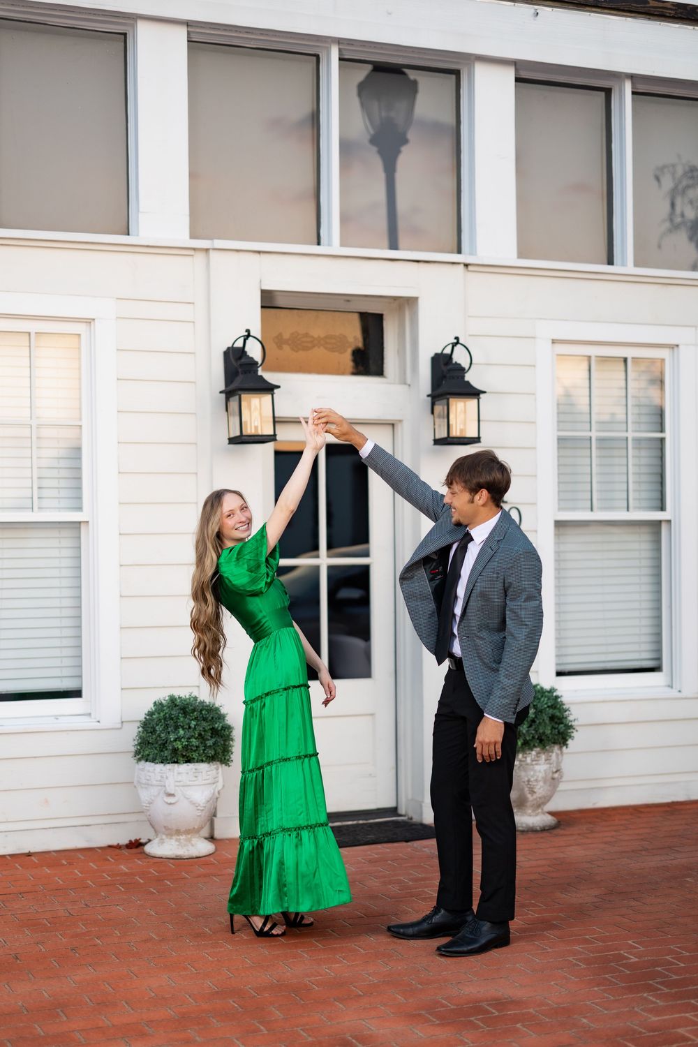 Couple dancing and twirling on brick patio in front of white building.