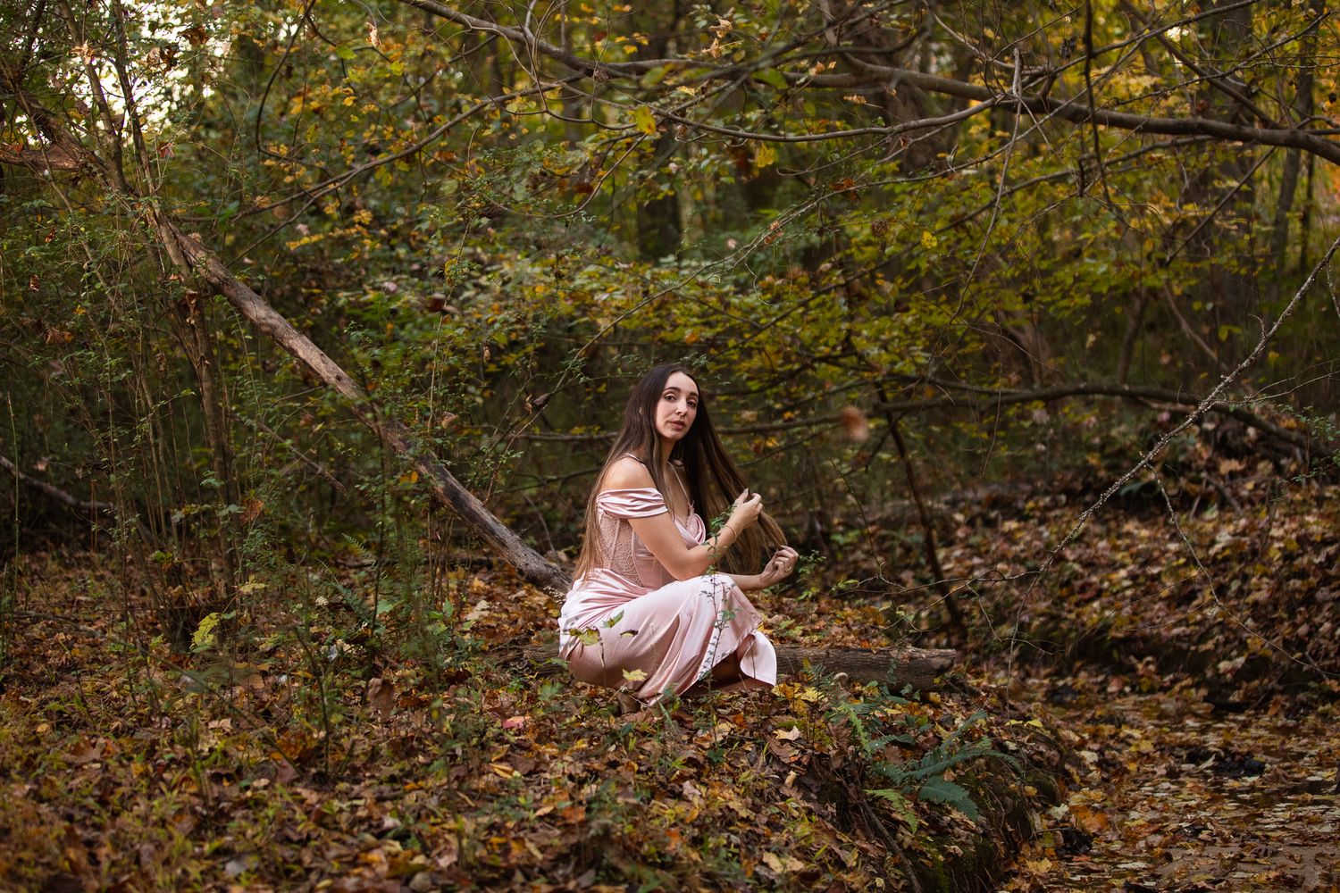 A person in a pink dress sits gracefully among autumn foliage in a wooded setting.