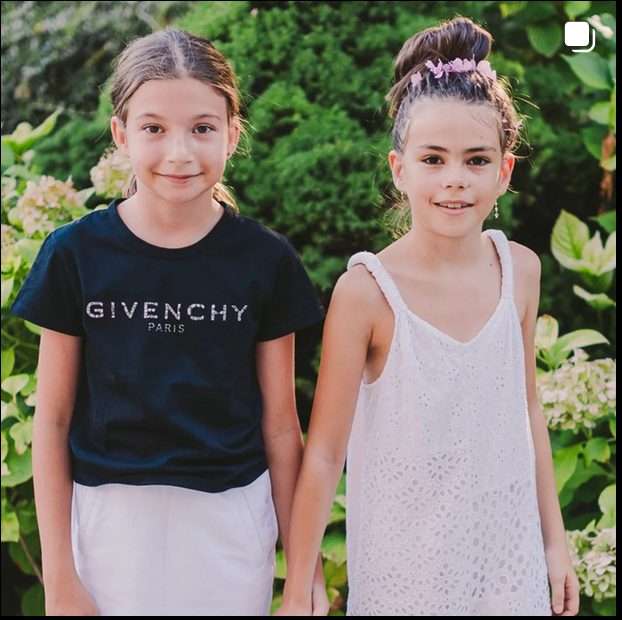 Children wearing summer outfits stand together against a green leafy background.