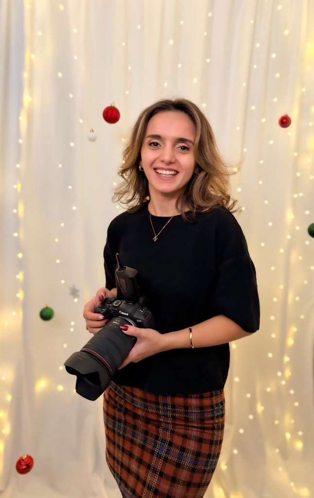 Person wearing black top and plaid skirt holds camera against backdrop of twinkling holiday lights.