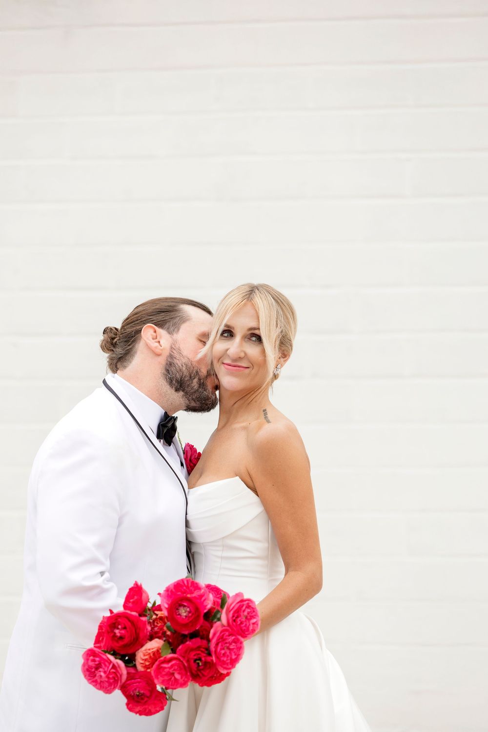 Romantic wedding couple embracing against white wall with vibrant pink roses.