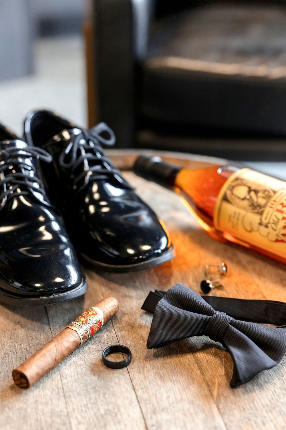 Black formal dress shoes with bow tie, cigar, and wedding ring arranged on wooden surface.