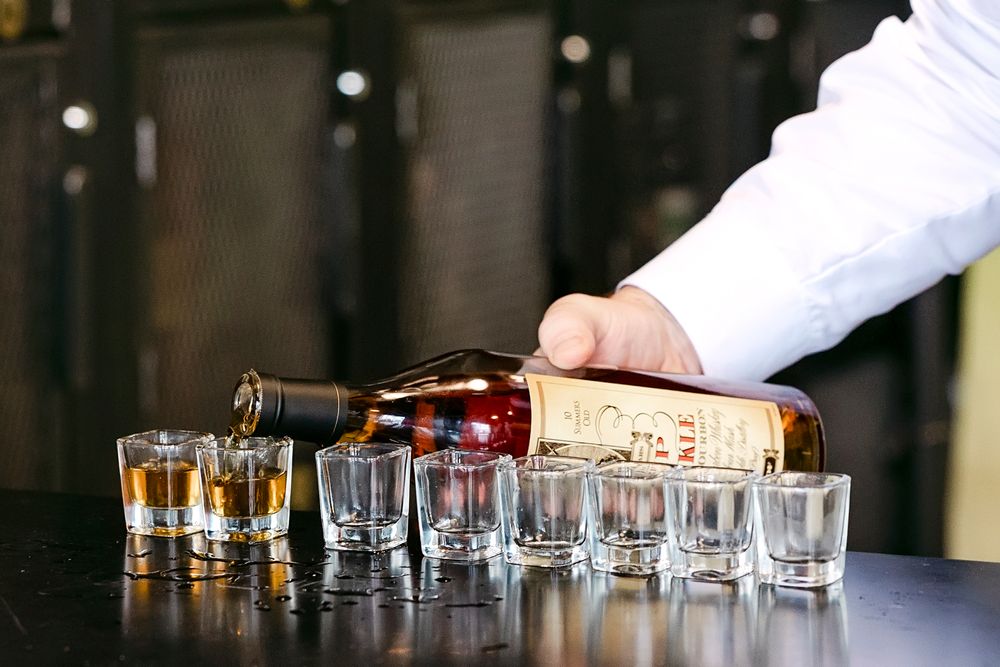 Row of empty shot glasses lined up on dark wooden bar counter with hands preparing drinks.