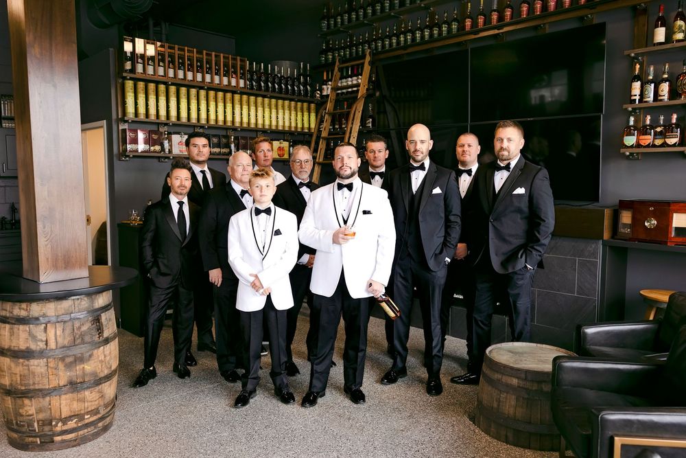 Wedding party members in black and white formal attire posing together in front of a rustic venue.