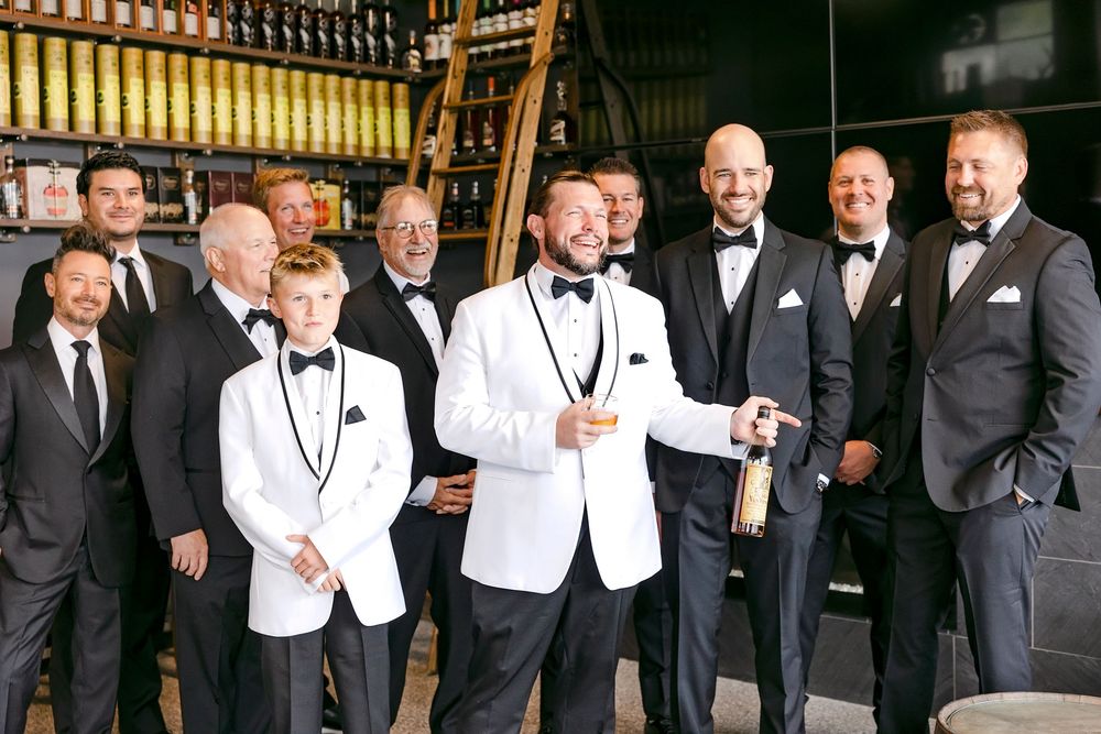 A group of groomsmen in black tuxedos and white suits pose together for formal wedding photos.