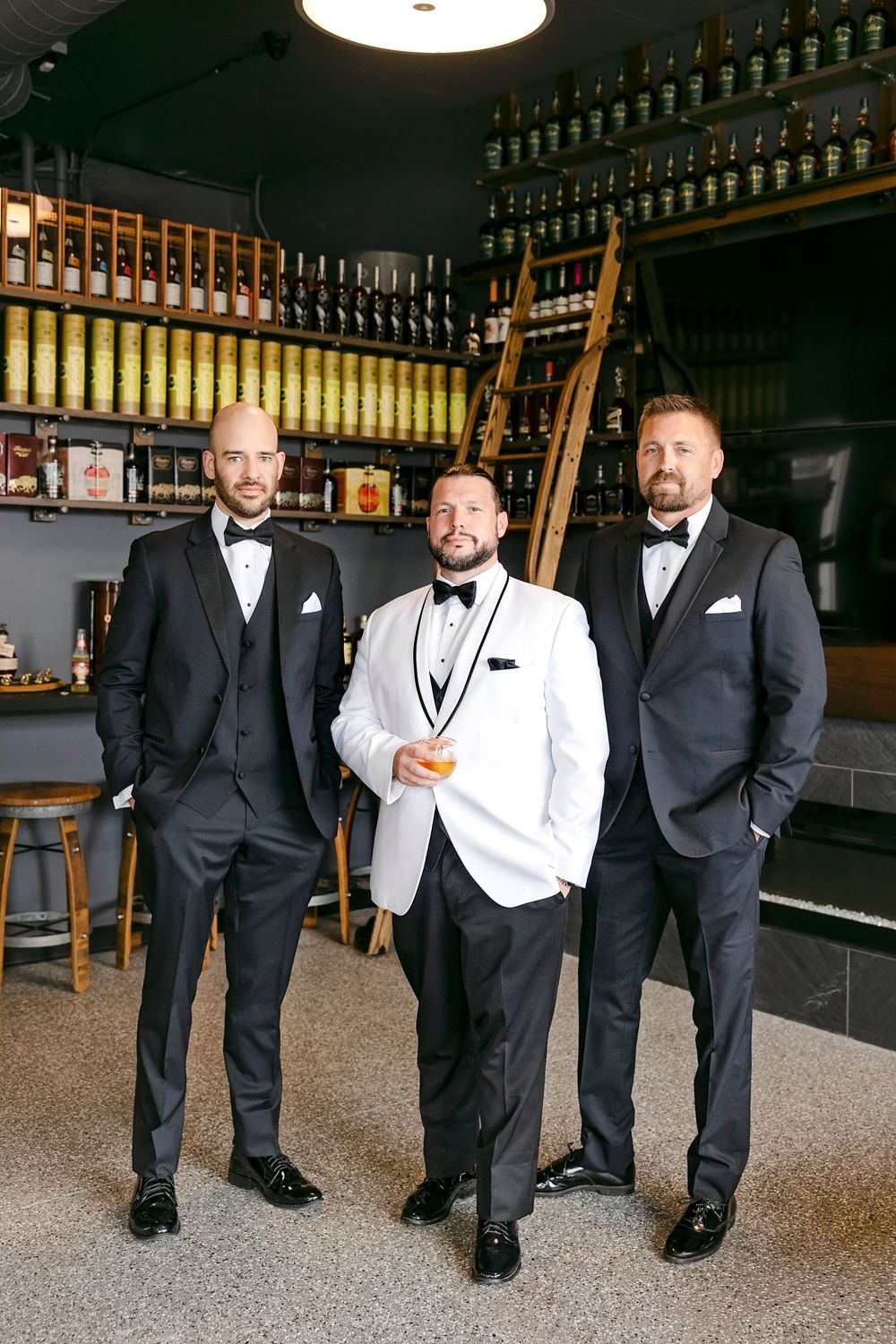 Groomsmen in black tuxedos and white vests stand together at a bar with bottles displayed in the background.
