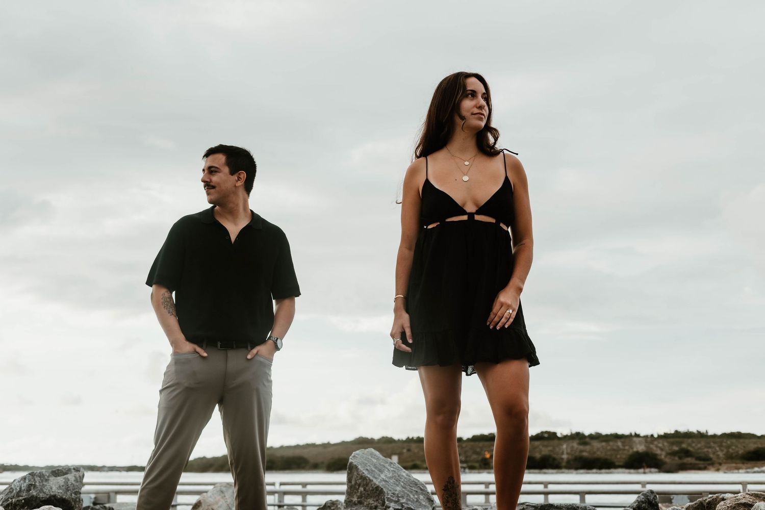 Couple in casual attire poses together on rocky coastline under cloudy sky.