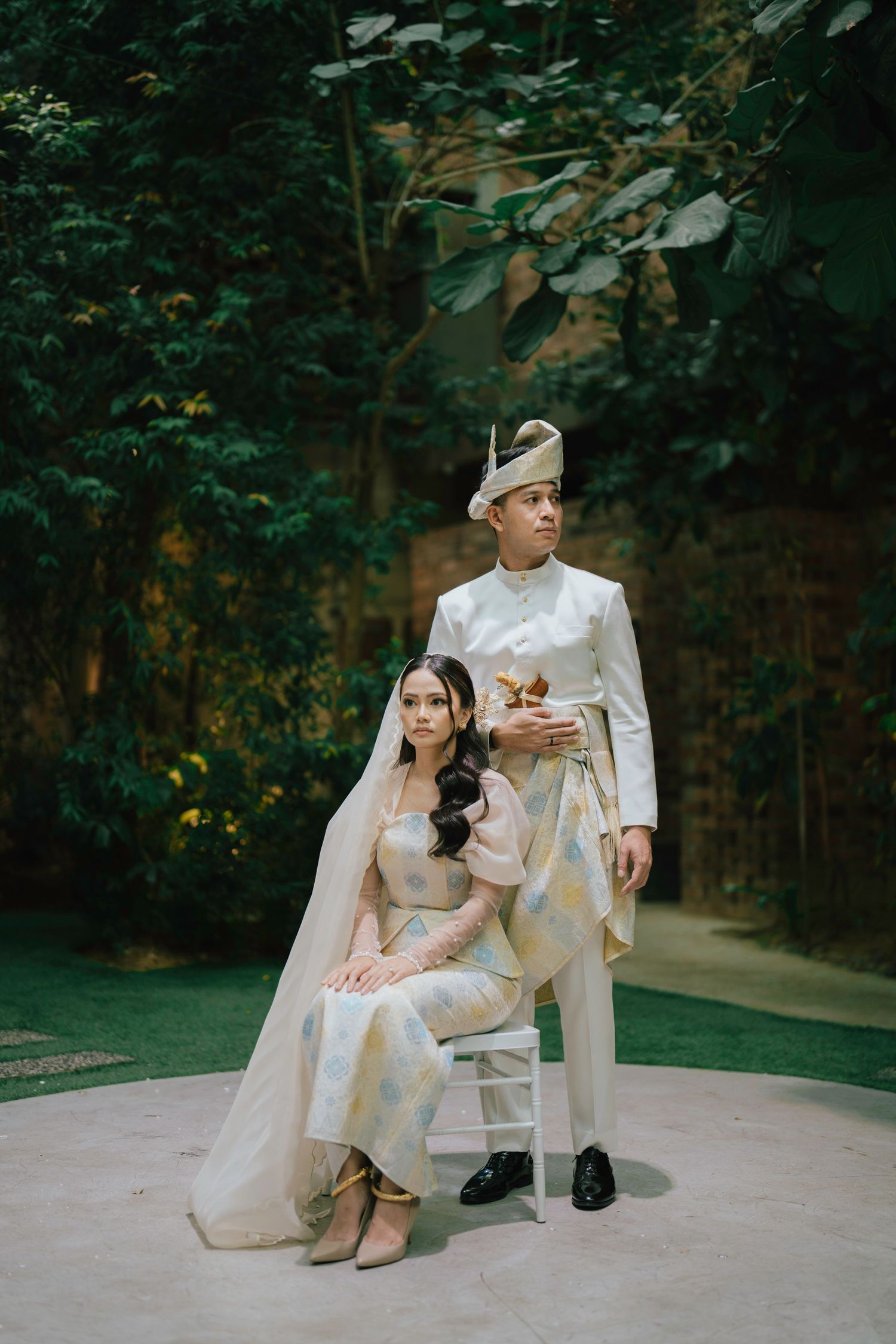 Malay couple in traditional white wedding attire pose elegantly in lush garden setting.