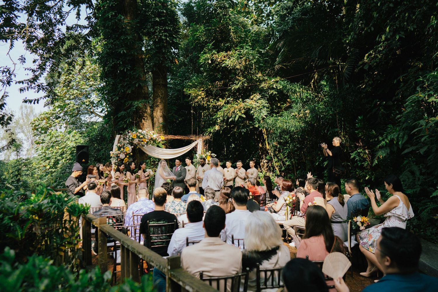 Outdoor wedding ceremony in lush garden setting with guests seated among tall trees and elegant decorations.