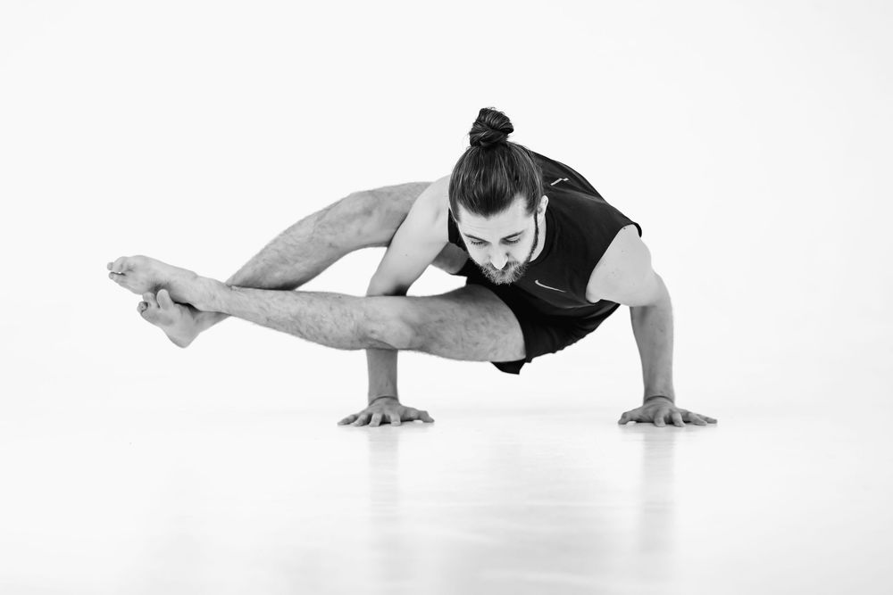 Dancer in black leotard performs advanced arm balance pose against white background.