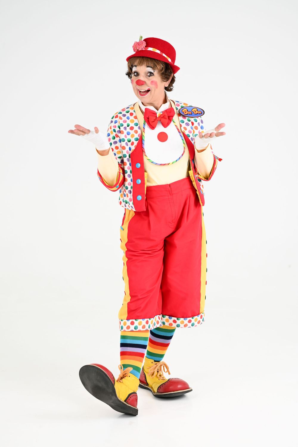 Clown in polka dot outfit with red overalls and striped socks poses with welcoming gesture.