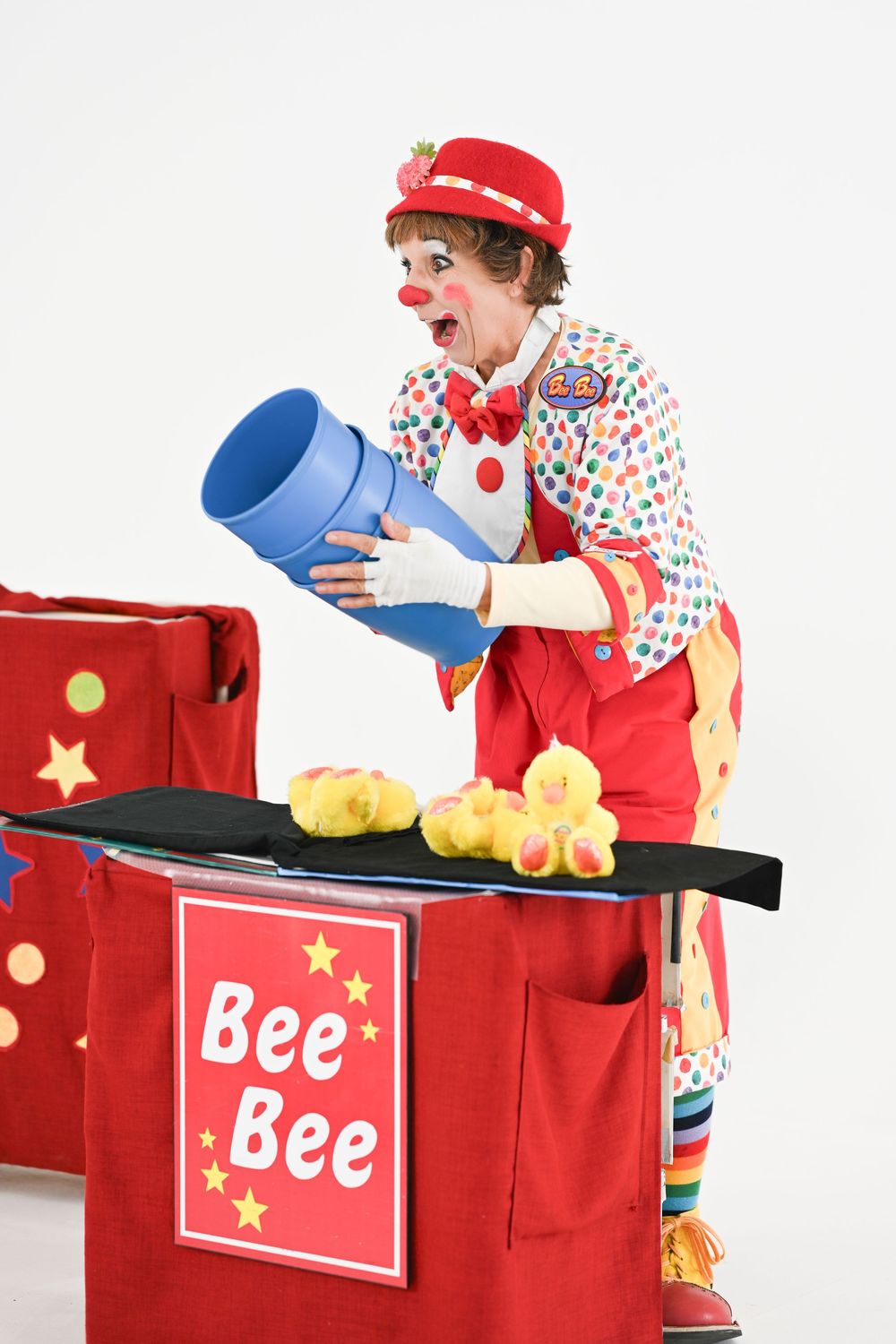 Colorful clown performer with polka dot outfit and red hat interacts with blue prop near red Bee Bee display cart.