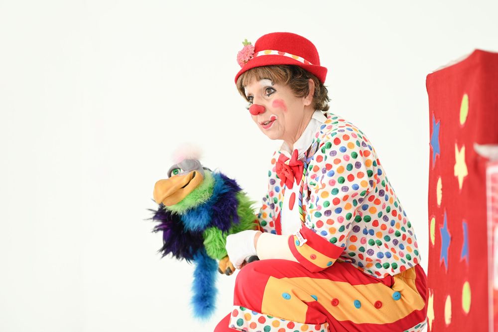 Clown performer with red hat holds colorful puppet while sitting next to red display.