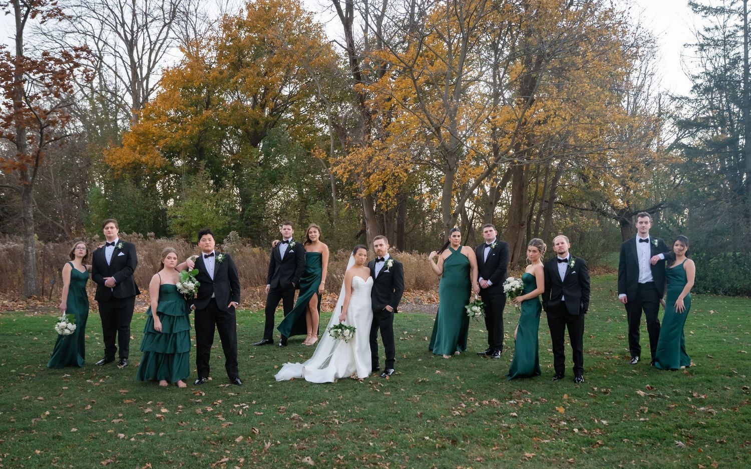 Wedding party stands together in a row on grass with autumn trees displaying vibrant fall foliage in the background.