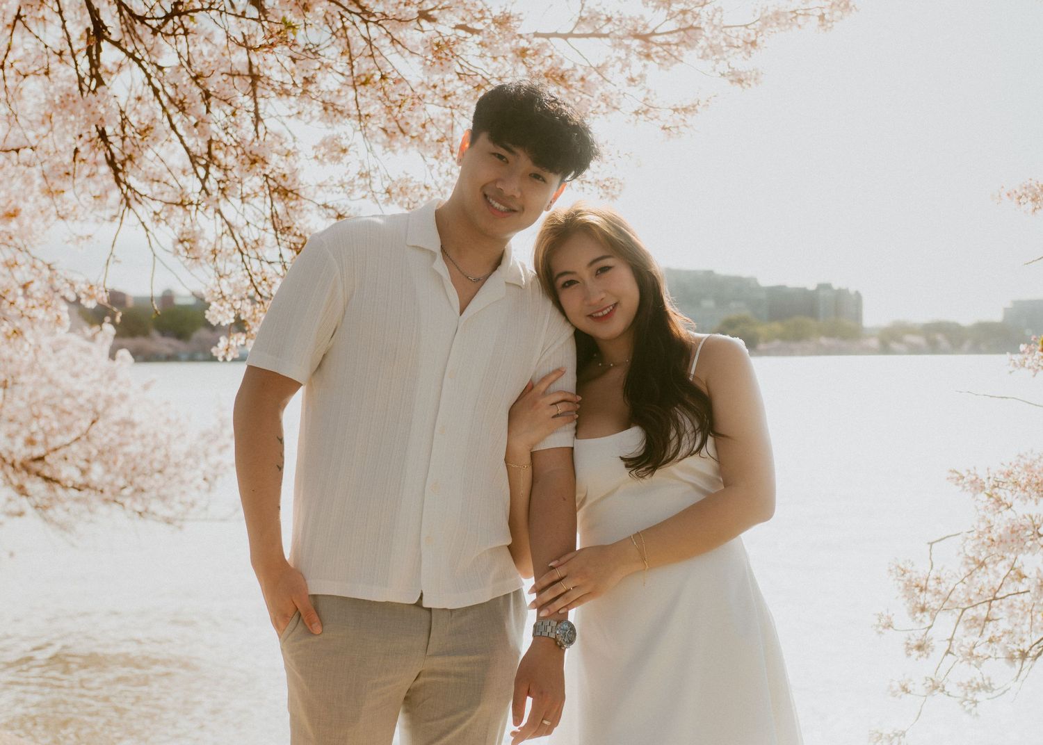 Couple in white outfits embracing under blooming cherry blossom trees during golden hour.