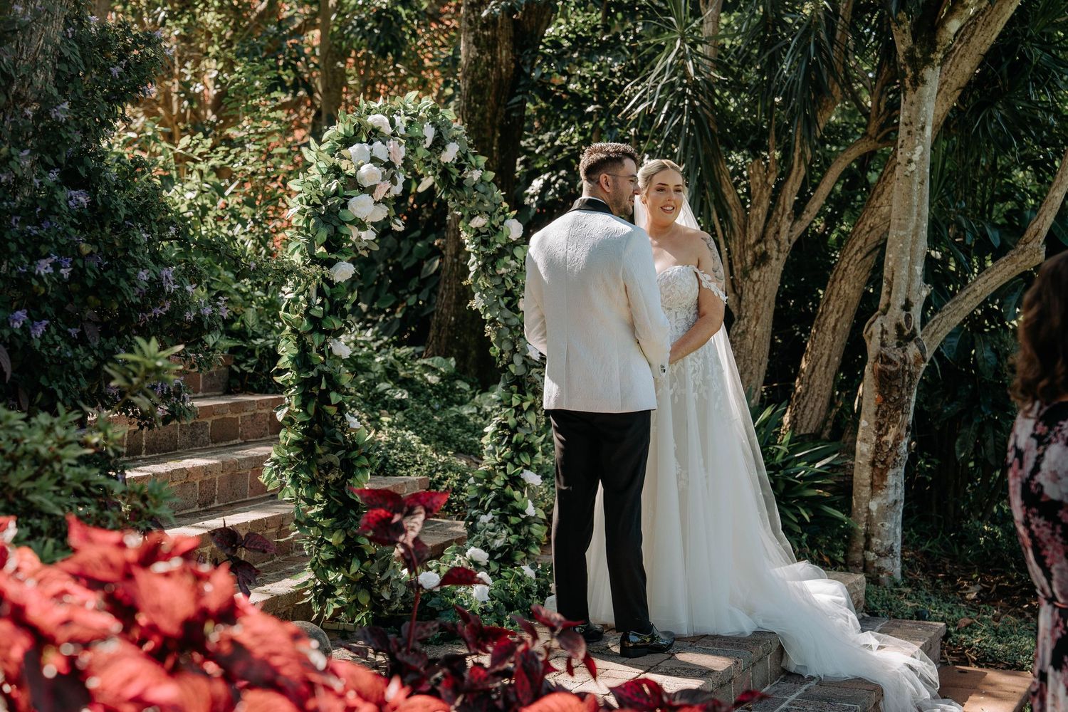 A couple shares a romantic moment in a garden setting with red flowers and lush greenery surrounding them.