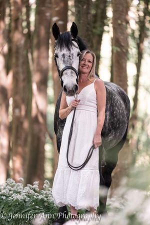 A dappled gray horse stands in a forest setting with someone in a white lace dress holding its lead rope.