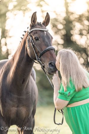 A dark brown horse and person in bright green dress share a tender moment in a sun-dappled outdoor setting.