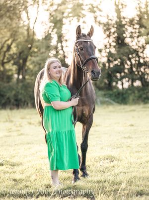 A woman in a bright green dress stands beside a dark brown horse in a sunlit grassy field during golden hour.