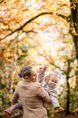 A parent in a beige coat playfully lifts and spins a small child wearing white during a golden autumn afternoon.