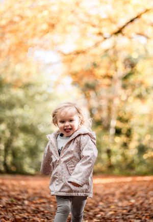 Child wearing a gray sweater plays joyfully in autumn leaves during a fall photoshoot in nature.