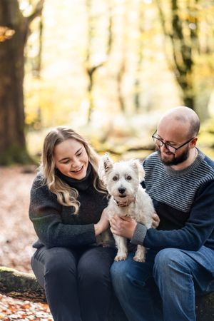 A couple sits outdoors on fallen autumn leaves with their small white dog in a series of candid photos.