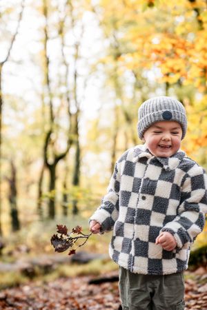 A small child in a checkered fleece jacket and gray beanie enjoys playing with fallen leaves on an autumn day.