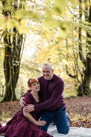 A couple shares joyful laughter while sitting on a blanket surrounded by autumn foliage in the woods.