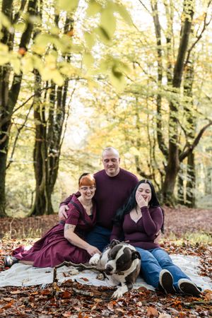 Three people relax with their Boston Terrier on colorful fallen leaves in an autumn forest setting.