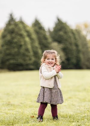 A young child in a cream sweater and plaid dress plays joyfully in a grassy field on an autumn day.