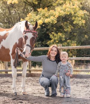A brown and white paint horse stands with people in a fenced paddock on an autumn day.